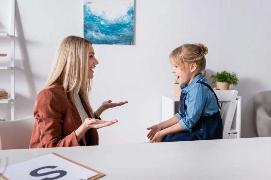 Side view of cheerful speech therapist talking with girl in consulting room.
